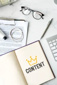 An open book with written content, a crown symbol on the heading, a pen, an extra worksheet, and a keyboard nearby – representing quality writing and strategy by content marketing services in Dubai UAE
